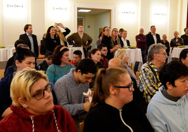 Eine Gruppe von Menschen, einige sitzend, andere stehend, besucht eine Ausstellung in einem hellen Raum. Einige Teilnehmer sind nach vorne gerichtet, während andere sich an hohen runden Tischen im Hintergrund aufhalten.