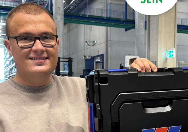 Eine lächelnde Person mit Brille und beigem Pullover hält eine Pappschachtel mit einem schwarzen Plastikbehälter darin. Oben rechts ist ein rundes Schild mit der Aufschrift lebenshilfe Wien #SICHTBAR SEIN in grüner Schrift zu sehen.