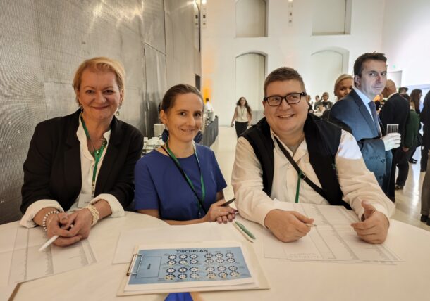 Drei Personen sitzen an einem Tisch mit Papieren und einem Sitzplan und lächeln in die Kamera. Sie tragen formelle Kleidung und Namensschilder. Im Hintergrund eines hellen, modernen Raums stehen andere Personen und unterhalten sich.