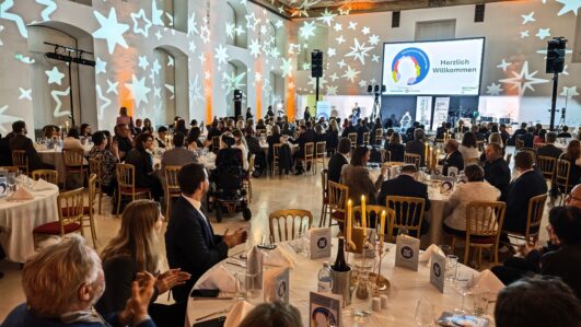 Ein großer, eleganter Bankettsaal, gefüllt mit Menschen, die an runden, mit Kerzen geschmückten Tischen sitzen. An die Wände sind sternförmige Lichter projiziert, und auf einer Leinwand an der Stirnseite steht 