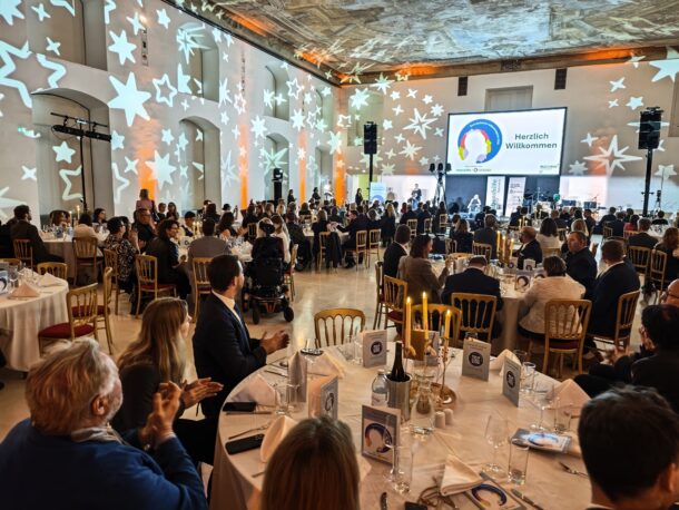 Ein großer, eleganter Bankettsaal, gefüllt mit Menschen, die an runden, mit Kerzen geschmückten Tischen sitzen. An die Wände sind sternförmige Lichter projiziert, und auf einer Leinwand an der Stirnseite steht 