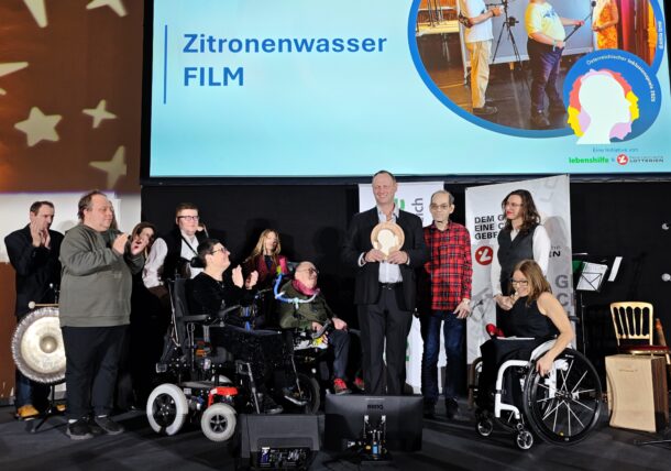 Eine Gruppe von Menschen, einige in Rollstühlen, posieren auf der Bühne und halten einen Preis. Auf einem Bildschirm hinter ihnen wird Zitronenwasser FILM mit Veranstaltungslogos angezeigt. Andere in der Gruppe applaudieren und lächeln.