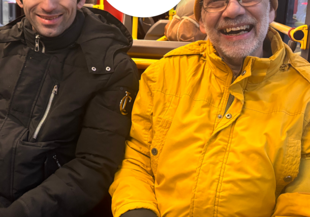 Zwei lächelnde Männer sitzen nebeneinander in einem Bus, beide tragen Winterjacken, eine schwarze und eine leuchtend gelbe. Über ihnen prangt ein kreisrundes Logo mit der Aufschrift 