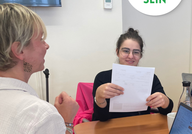 Zwei Frauen sitzen an einem Tisch mit Dokumenten und einem Laptop. Eine Frau lächelt und hält ein Papier hoch, die andere schaut ihr zu. Auf einem runden Schild steht in deutscher Sprache 