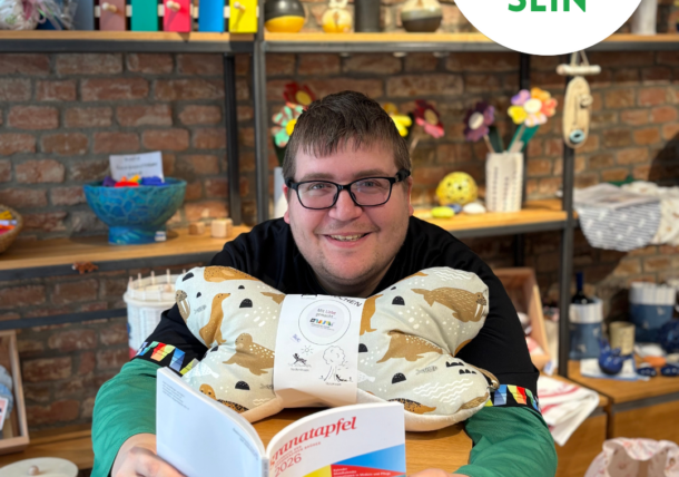 Ein lächelnder Mann mit Brille hält ein Buch und einen Plüschhund in einem bunten Laden. Hinter ihm stehen Regale mit Dekorationsartikeln. Auf einem runden Schild steht in grüner Schrift Lebenshilfe Wien #SICHTBAR SEIN.