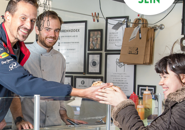 Ein lächelnder Verkäufer reicht einer Frau an einer Essensausgabe ein Sandwich, während ein anderer Mitarbeiter hinter der Theke steht. Die Szene ist fröhlich, darüber steht ein Schild mit der Aufschrift lebenshilfe Wien #SICHTBAR SEIN.
