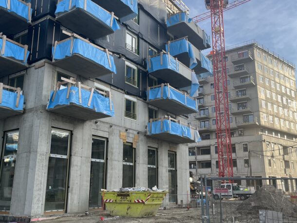 Ein modernes, im Bau befindliches Wohngebäude mit blauen Balkonverkleidungen, einem gelben Müllcontainer und Kränen im Hintergrund. Im Vordergrund sind Baumaterialien und Zäune zu sehen.