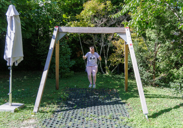 Eine Person, die ein weißes T-Shirt und rosa Shorts trägt, sitzt auf einer Schaukel in einem grünen Garten, umgeben von Bäumen und Schatten. Links neben der Schaukel steht ein ungeöffneter weißer Regenschirm.
