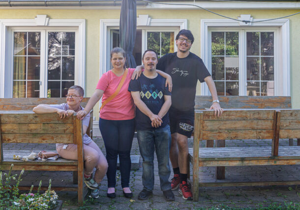 Vier Erwachsene posieren fröhlich draußen vor einem hellgelben Gebäude mit großen Fenstern. Zwei stehen und lächeln, die Arme umeinander gelegt, während die anderen beiden sitzen und sich auf Holzbänke stützen.