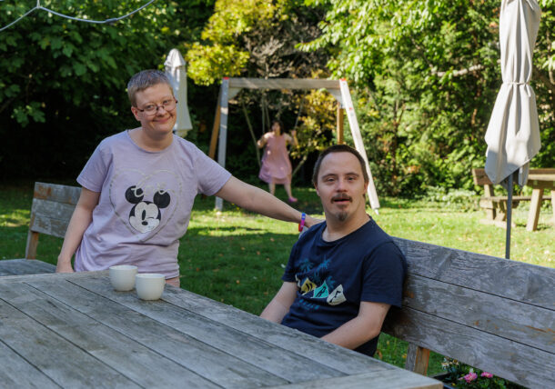 Zwei Erwachsene mit Down-Syndrom sitzen in einem sonnigen Garten an einem hölzernen Picknicktisch und genießen die Zeit im Freien. Der eine steht und lächelt, während der andere sitzt. Im Hintergrund spielt ein Kind auf einer Schaukel.
