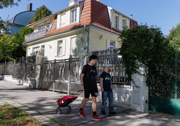 Zwei Personen gehen auf einem sonnigen Bürgersteig vor einem gelben Haus mit rotem Dach. Eine Person zieht einen kleinen roten Wagen, und beide scheinen sich zu unterhalten, während sie an Grünpflanzen und einem Zaun vorbeigehen.