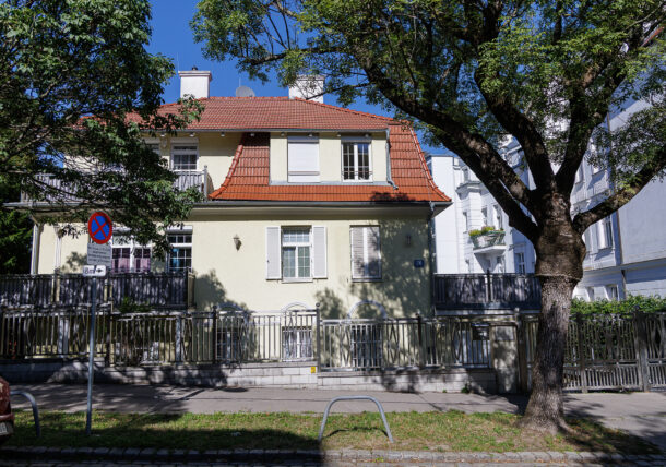 Ein zweistöckiges cremefarbenes Haus mit rotem Ziegeldach und weißen Fensterläden, das von einem Metallzaun umgeben ist und an einem sonnigen Tag von großen Bäumen beschattet wird. Auf dem Gehweg davor ist ein Parkverbotsschild zu sehen.