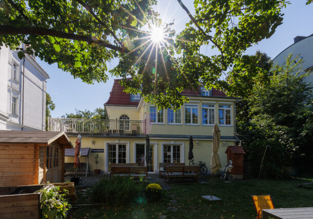 Das Sonnenlicht scheint durch die Blätter eines großen Baumes in einem Garten und beleuchtet ein gelbes zweistöckiges Haus mit roten Dachziegeln. In dem grünen Hof stehen Holzbänke, ein Schuppen und geschlossene Sonnenschirme.