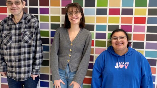 Drei Personen stehen lächelnd vor einer bunten Mosaikwand. Die Person links trägt eine Brille und ein kariertes Hemd, die mittlere Person eine graue Strickjacke und die Person rechts eine Brille und einen blauen Kapuzenpullover.