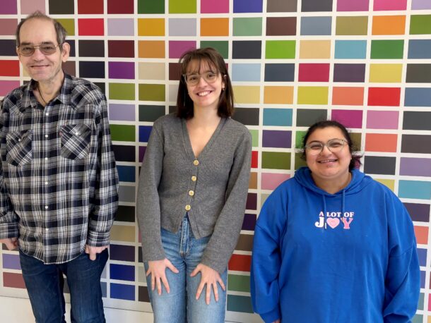 Drei Personen stehen lächelnd vor einer bunten Mosaikwand. Die Person links trägt eine Brille und ein kariertes Hemd, die mittlere Person eine graue Strickjacke und die Person rechts eine Brille und einen blauen Kapuzenpullover.