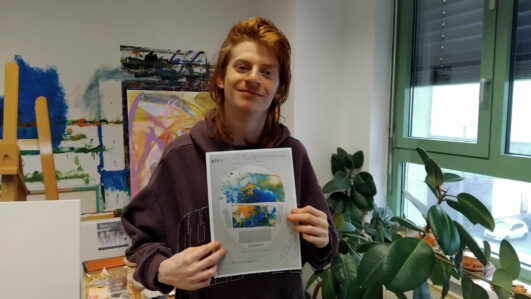 Eine Person mit langen rötlichen Haaren, die einen dunklen Kapuzenpulli trägt, hält lächelnd ein bedrucktes Papier mit einer bunten Landkarte hoch. Sie stehen in einem Raum mit Kunstzubehör, Pflanzen und einem großen Fenster.