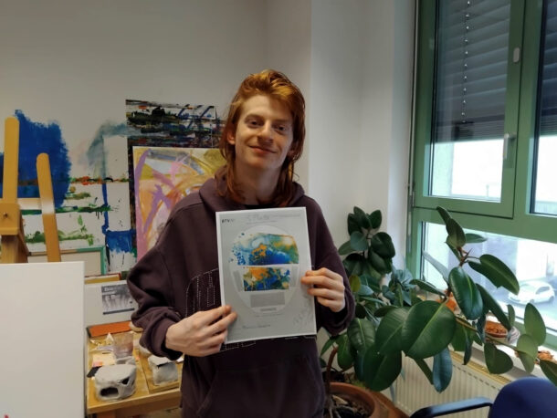 Eine Person mit langen rötlichen Haaren, die einen dunklen Kapuzenpulli trägt, hält lächelnd ein bedrucktes Papier mit einer bunten Landkarte hoch. Sie stehen in einem Raum mit Kunstzubehör, Pflanzen und einem großen Fenster.