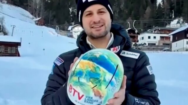 Ein Mann in Wintersportkleidung steht im Schnee, lächelt und hält einen Skihelm, der mit einer bunten Weltkarte und Logos verziert ist. Im Hintergrund sind verschneite Gebäude und Bäume zu sehen.