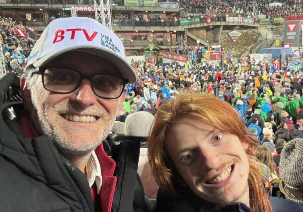 Zwei Personen lächeln für ein Selfie bei einer nächtlichen Sportveranstaltung im Freien, mit Stadionbestuhlung und einem großen, hell erleuchteten Publikum im Hintergrund. Eine Person trägt eine Brille und einen weißen Hut, die andere hat lange rote Haare.