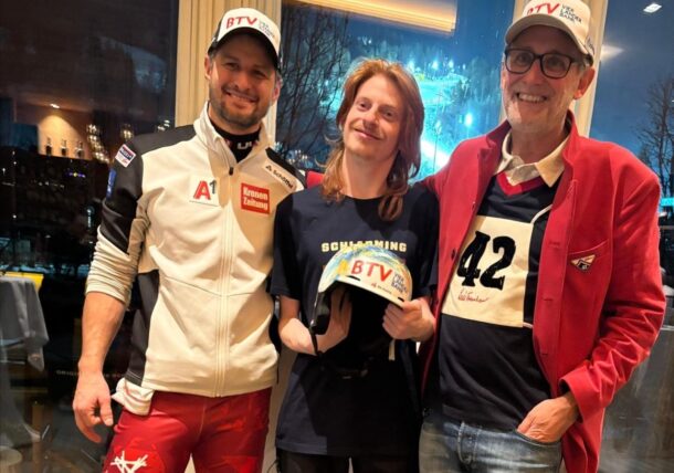 Drei Personen stehen lächelnd in einem Haus. Die Person auf der linken Seite trägt ein sportliches Outfit, die mittlere Person hält einen Helm, und die Person auf der rechten Seite trägt eine Brille, einen roten Blazer und eine Startnummer. Durch das Fenster hinter ihnen ist eine nächtliche Stadtlandschaft zu sehen.