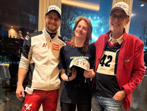 Drei Personen stehen lächelnd in einem Haus. Die Person auf der linken Seite trägt ein sportliches Outfit, die mittlere Person hält einen Helm, und die Person auf der rechten Seite trägt eine Brille, einen roten Blazer und eine Startnummer. Durch das Fenster hinter ihnen ist eine nächtliche Stadtlandschaft zu sehen.