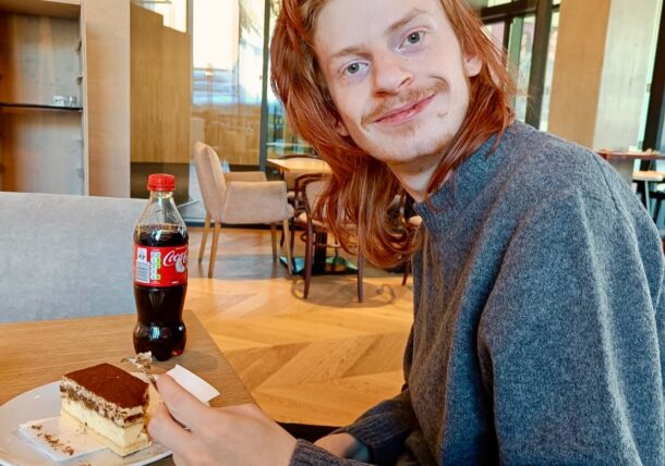 Eine Person mit langen roten Haaren und einem Schnurrbart, die einen grauen Pullover trägt, sitzt lächelnd an einem Cafétisch und hält eine Gabel mit Tiramisu-Kuchen und eine Flasche Coca-Cola vor sich. Das Café hat einen Holzfußboden und ist modern eingerichtet.