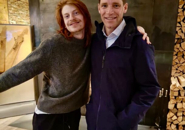 Zwei Personen stehen in einem Haus und lächeln in die Kamera. Die eine hat langes rotes Haar und einen grauen Pullover. Sie hat den Arm um die andere gelegt, die kurze braune Haare hat und einen marineblauen Mantel trägt. Hinter ihnen steht ein moderner Kamin mit gestapeltem Brennholz.