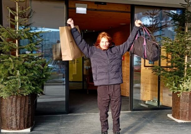 Eine Person in dunkler Jacke und Hose steht lächelnd mit erhobenen Armen, in der einen Hand eine Einkaufstasche, in der anderen einen Seesack, vor einem mit immergrünen Topfbäumen geschmückten Hauseingang.