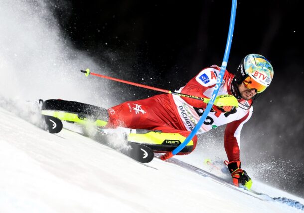 Ein rot-weiß gekleideter Skifahrer mit Helm und Skibrille lehnt sich stark vor, während er auf einer verschneiten Piste durch ein Slalomtor rast und dabei eine Schneeschleuder aufwirbelt.