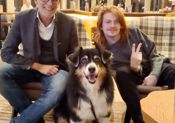 Zwei Menschen sitzen in einem Haus auf Stühlen neben einem großen schwarz-weißen Hund. Eine Person lächelt, die andere macht ein Friedenszeichen. Der Hintergrund zeigt warme Beleuchtung und modernes Dekor.