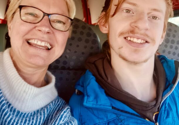 Eine lächelnde Frau mit Brille und einem hellblauen Pullover sitzt neben einem lächelnden jungen Mann in einer blauen Jacke. Sie sitzen dicht beieinander in einem Fahrzeug mit gemusterten Sitzen.