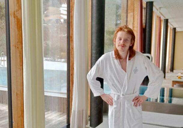 Eine Person mit langem rotem Haar und Schnurrbart steht in einem weißen Bademantel, die Hände in die Hüften gestemmt, vor bodentiefen Fenstern, während draußen der Pool und die verschneite Landschaft zu sehen sind.