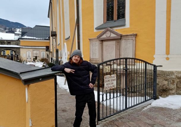 Eine Person in Winterkleidung lehnt an einem gelben Gebäude mit gewölbten Fenstern und einem Tor. Auf dem Boden und den nahen Dächern liegt Schnee, und im Hintergrund sind Berge zu sehen.