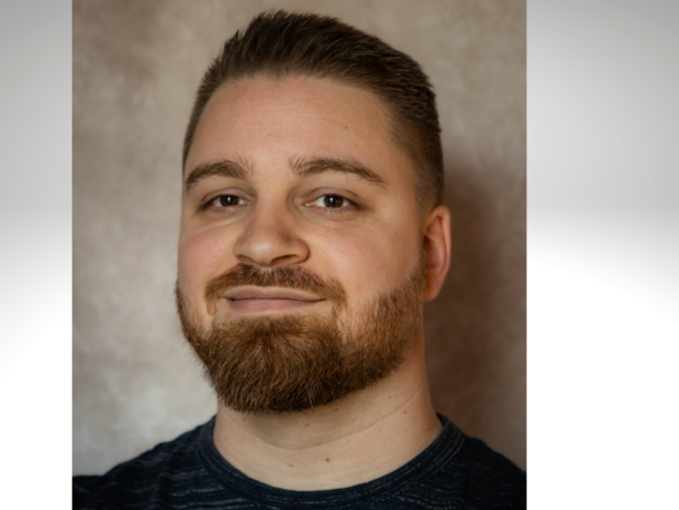 Ein Mann mit kurzen braunen Haaren und einem Vollbart lächelt leicht in die Kamera, er trägt ein dunkles Hemd. Der Hintergrund ist neutral und sanft verschwommen.