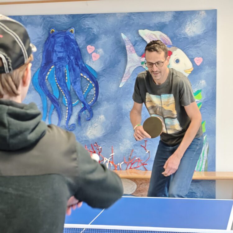 Zwei Personen spielen in einer Halle Tischtennis. Der eine, mit Mütze und dunkler Jacke, schaut in die Kamera, der andere, mit Brille und T-Shirt, ist dabei, den Ball zu schlagen. Ein farbenfrohes Wandgemälde zum Thema Meer schmückt die Wand hinter ihnen.