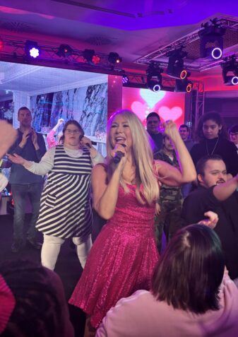 Eine Frau in einem glitzernden rosafarbenen Kleid singt auf einer farbenfrohen Bühne enthusiastisch in ein Mikrofon, umgeben von jubelnden Menschen, die tanzen und ihre Arme in die Höhe recken, während die Lichter leuchten.