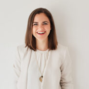 Eine Frau mit glattem braunem Haar, die einen weißen Blazer und eine lange Halskette trägt, steht lächelnd vor einem schlichten weißen Hintergrund.