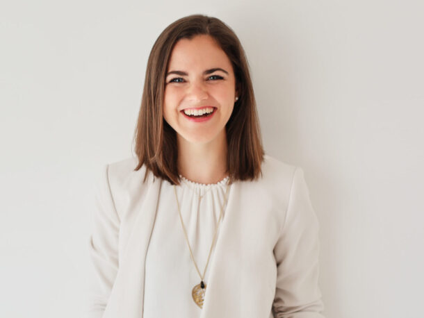 Eine Frau mit glattem braunem Haar, die einen weißen Blazer und eine lange Halskette trägt, steht lächelnd vor einem schlichten weißen Hintergrund.