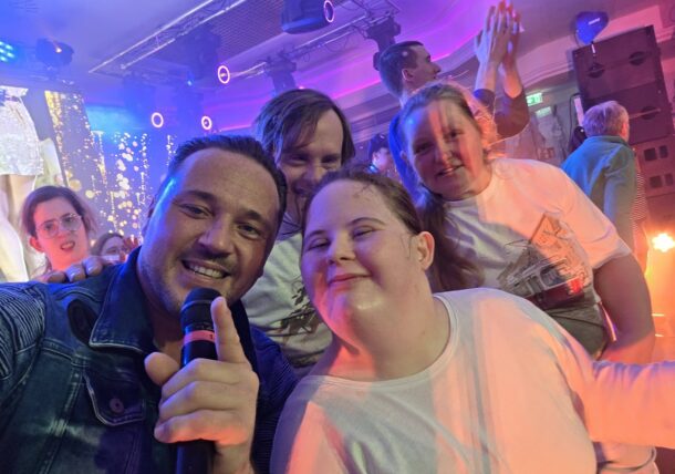 Eine Gruppe von Menschen lächelt und posiert für ein Selfie bei einer lebhaften Veranstaltung. Eine Person im Vordergrund hält ein Mikrofon, während im Hintergrund bunte Lichter und jubelnde Zuschauer zu sehen sind.