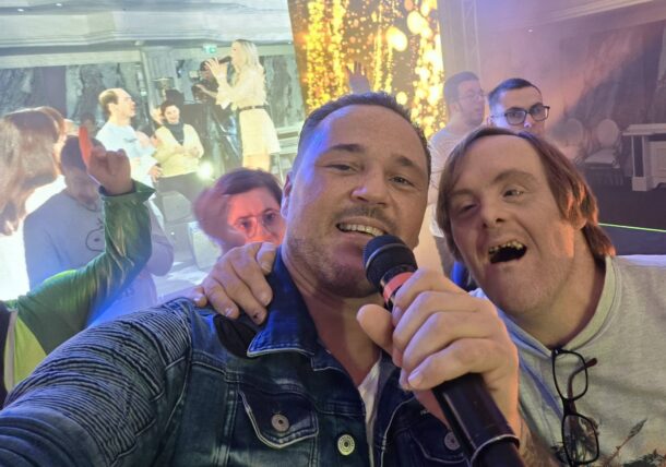 Eine Gruppe von Menschen lächelt und posiert für ein Selfie bei einer lebhaften Veranstaltung. Ein Mann im Vordergrund hält ein Mikrofon, und ein anderer Mann mit Brille lehnt sich hinein. Im Hintergrund sind helle Lichter und festliche Dekorationen zu sehen.