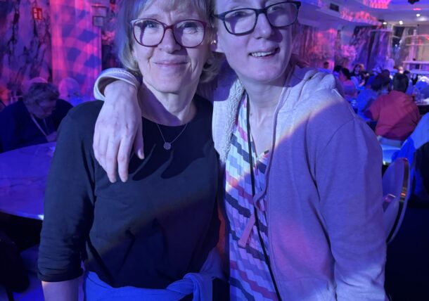 Zwei Brillenträger lächeln und posieren bei einer Indoor-Veranstaltung unter strahlend blauem und violettem Licht zusammen, wobei eine Person den Arm um die Schultern der anderen legt. Andere Gäste sitzen an Tischen im Hintergrund.