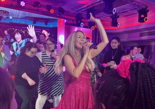 Eine Frau in einem glitzernden rosa Kleid singt auf der Bühne in ein Mikrofon, umgeben von einer begeisterten Menge tanzender und lächelnder Menschen unter bunten Bühnenlichtern.