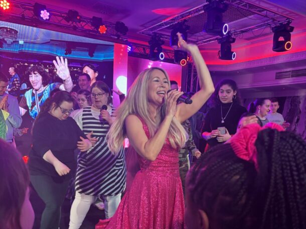 Eine Frau in einem glitzernden rosa Kleid singt auf der Bühne in ein Mikrofon, umgeben von einer begeisterten Menge tanzender und lächelnder Menschen unter bunten Bühnenlichtern.