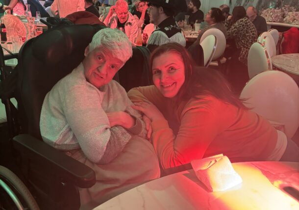 Zwei Frauen, die eine im Rollstuhl, die andere neben ihr kauernd, lächeln in die Kamera in einem belebten, eleganten Restaurant mit Marmortischen und -wänden. Die warme rote und grüne Beleuchtung verleiht der Szene eine festliche Schlagerfest-Atmosphäre.