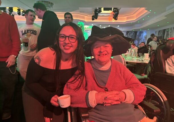 Eine lächelnde junge Frau mit Brille posiert neben einer älteren Frau im Rollstuhl, die einen großen schwarzen Hut trägt. Beide wirken glücklich bei einer lebhaften Veranstaltung in einer Halle mit Menschen und Tischen im Hintergrund.