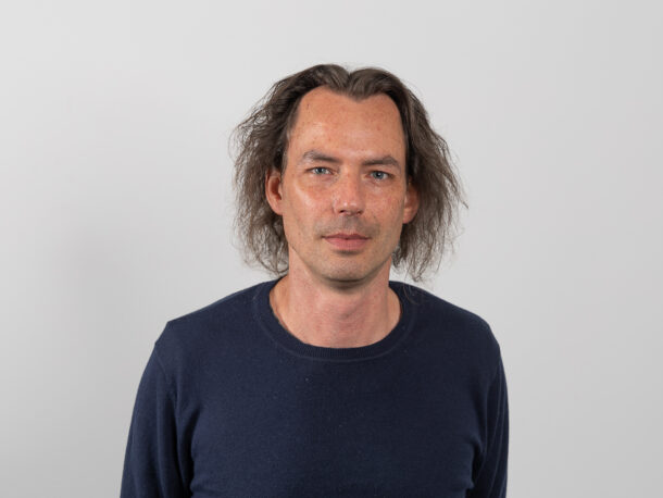 Ein Mann mit gewelltem, schulterlangem braunem Haar und einem dunkelblauen Pullover steht vor einem schlichten hellgrauen Hintergrund und schaut mit neutralem Blick direkt in die Kamera.
