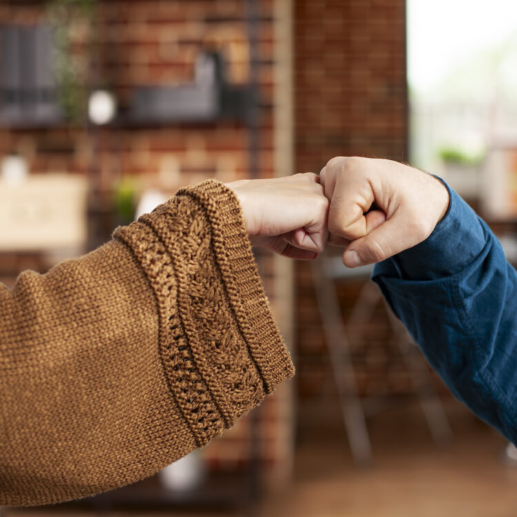 Zwei Personen geben sich in einem Haus die Hand, die eine in einem braunen Strickpullover, die andere in einem blauen Hemd, das Schutz vor Gewalt symbolisiert, vor dem unscharfen Hintergrund eines Zimmers mit Backsteinwänden.