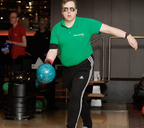 Eine Person, die ein grünes Hemd, eine schwarze Hose und orangefarbene Schuhe trägt, ist beim Bowling und hält eine blaue Bowlingkugel mitten im Schwung auf einer Bowlingbahn. Im Hintergrund sind andere Personen und Bowlingkugeln zu sehen.
