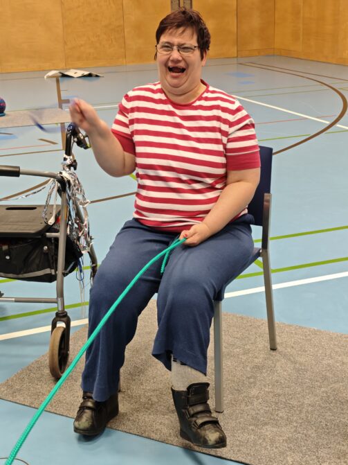 Eine Frau mit Brille, einem rot-weiß gestreiften Hemd und einer blauen Hose sitzt auf einem Stuhl in einer Turnhalle. Sie lächelt, während sie ein grünes Seil in der Hand hält und eine Gehhilfe neben sich hat.