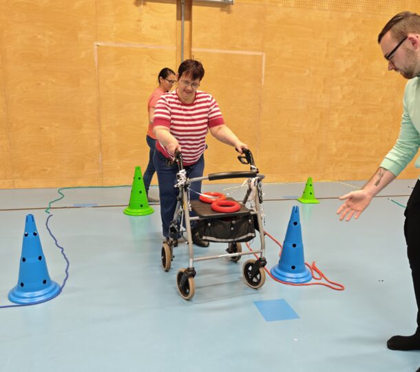 Eine Frau, die einen Rollator benutzt, navigiert um blaue und grüne Hütchen in einer Turnhalle, während ein Mann in der Nähe unterstützend gestikuliert und eine weitere Person hinter ihr steht.